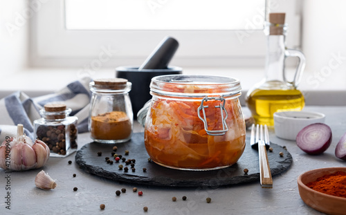 Spicy Korean appetizer kimchi made of cabbage with carrots in a glass jar with spices garlic and onions on a dark dish. Horizontal orientation, gray background, no people.