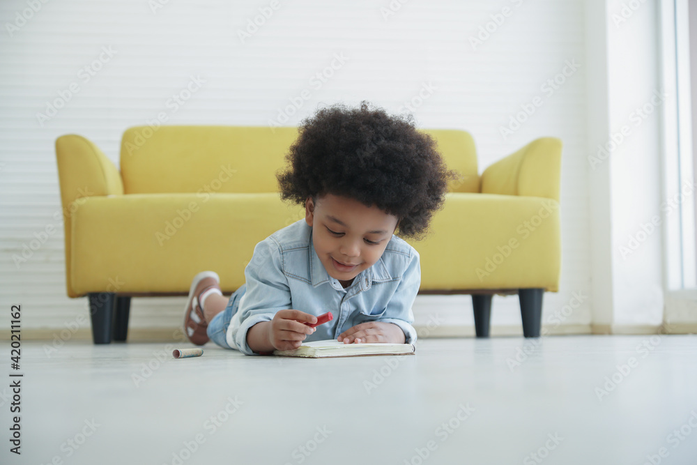 Little African boy holding red crayon in hand drawing coloring on book and lying on the floor at home
