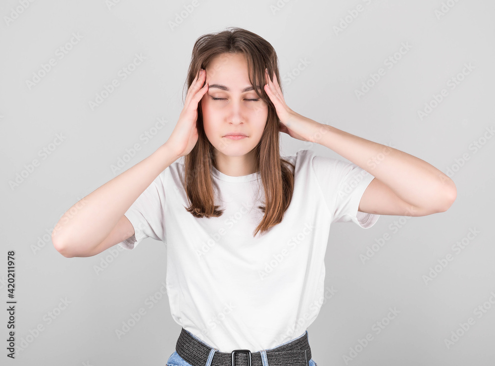 Young brunette in white t-shirt and jishes on isolated gray background suffering from headache in despair and stress due to pain and migraine. Hands on the head. Young brunette in white t-shirt and jishes on isolated gray background suffering from headache in despair and stress due to pain and migraine. Hands on the head.