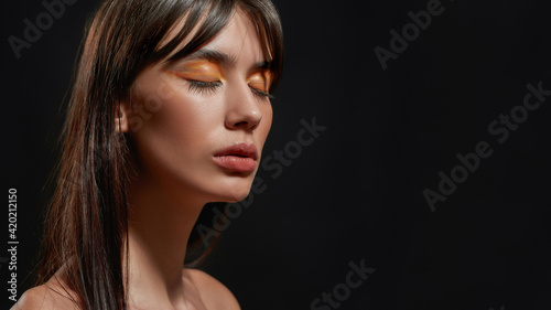 Close up beauty portrait of young brunette woman with colorful orange makeup posing with eyes closed isolated over black background