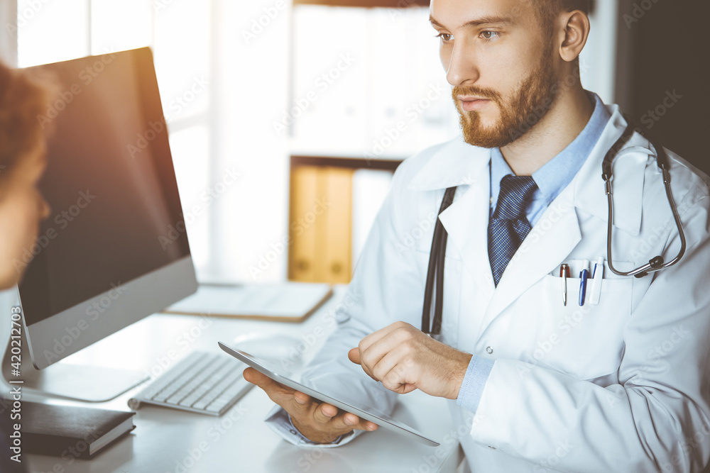 Friendly red-bearded doctor and patient woman discussing current health examination while sitting and using tablet computer in sunny clinic