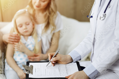 Doctor and patient. Pediatrician using clipboard while examining little girl with her mother at home. Sick and unhappy child at medical exam