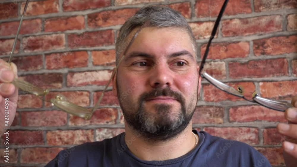 A bearded man tries on different glasses. Short haircut. Blue T-shirt. Poor eyesight. Choice of glasses. Optics. Against the background of a brick wall. Large portrait. A bearded man tries on different glasses. Short haircut. Blue T-shirt. Poor eyesight. Choice of glasses. Optics. Against the background of a brick wall. Large portrait.