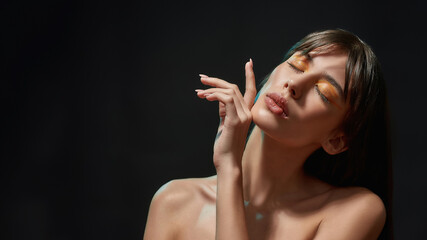 Beauty portrait of young brunette woman with healthy brown hair and professional orange makeup touching her chin, posing with eyes closed isolated over black background