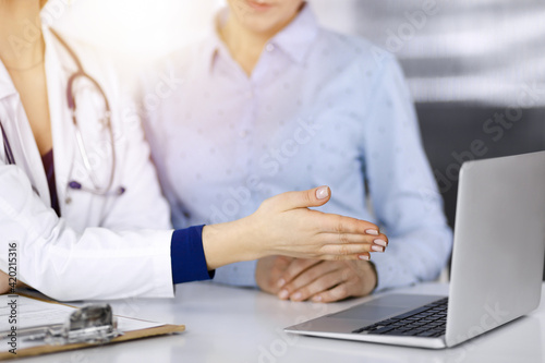 Unknown woman- doctor and her patient are discussing patient s blood test  while sitting together at the desk in the sunny cabinet in a clinic. Female physician  with a stethoscope  is using a laptop