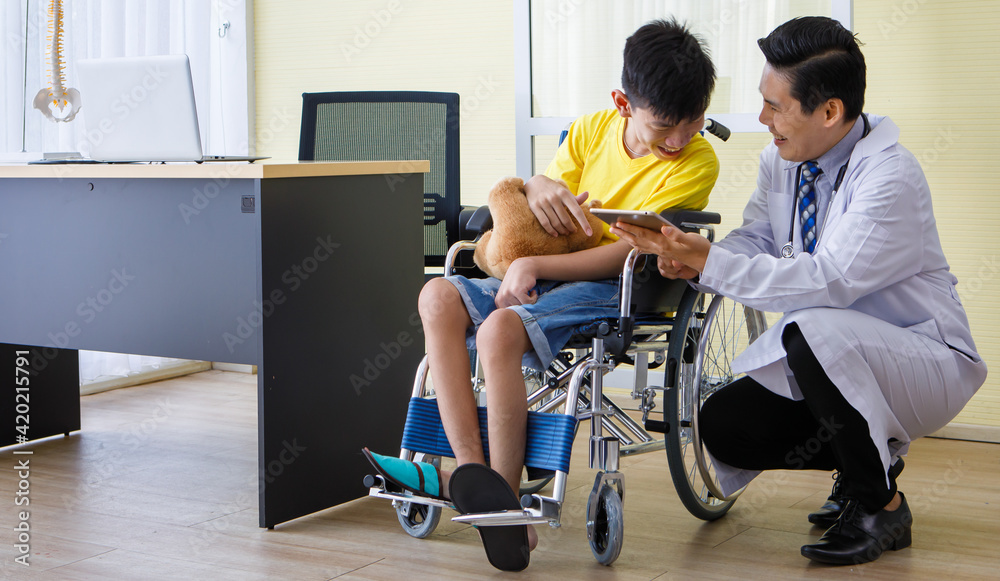 Asian doctor talking, support, and encouragement to a young disabled boy in a wheelchair. He shows tablet computer screen to the patient Asian doctor talking, support, and encouragement to a young disabled boy in a wheelchair. He shows tablet computer screen to the patient