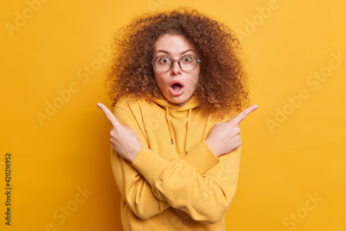 Shocked terrified emotive woman with curly hair points sideways at variants indicates right and left keeps mouth wide opened dressed in hoodie cannot choose between two items isolated on yellow wall © wayhome.studio 