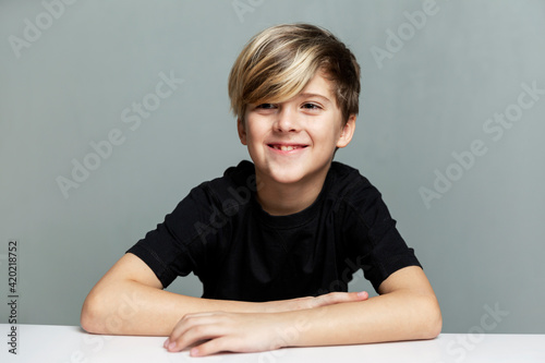 A boy in a black T-shirt with a fashionable hairstyle sits at a table and laughs. Grey background.