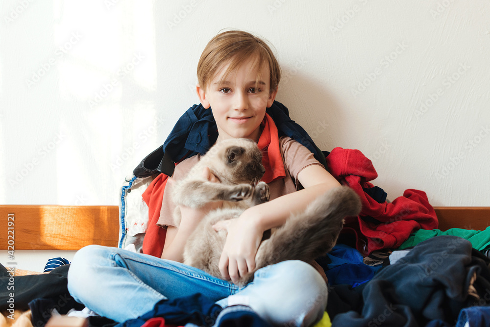 Cute boy plays with cat. Untidy boy sitting on the bed full of his dirty clothes. Home chores housework. Cute boy plays with cat. Untidy boy sitting on the bed full of his dirty clothes. Home chores housework.