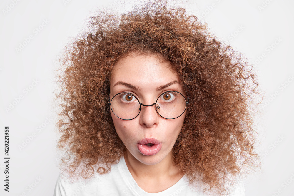 Headshot of surprised European woman with curly bushy hair looks with amazement at camera cannot believe in shocking news wears round spectacles isolated over white studio background. Omg concept Headshot of surprised European woman with curly bushy hair looks with amazement at camera cannot believe in shocking news wears round spectacles isolated over white studio background. Omg concept