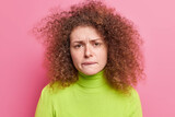 Photo of frustrated unhappy woman with curly hair bites lips looks nervously at camera feels anxious dressed in casual green turtleneck isolated over pink background. Perplexed troubled female