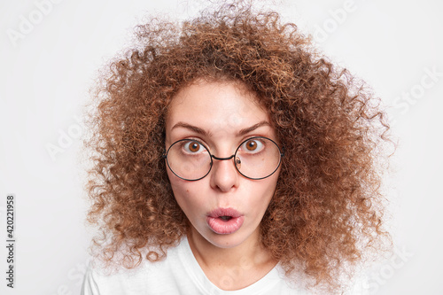 Headshot of surprised European woman with curly bushy hair looks with amazement at camera cannot believe in shocking news wears round spectacles isolated over white studio background. Omg concept