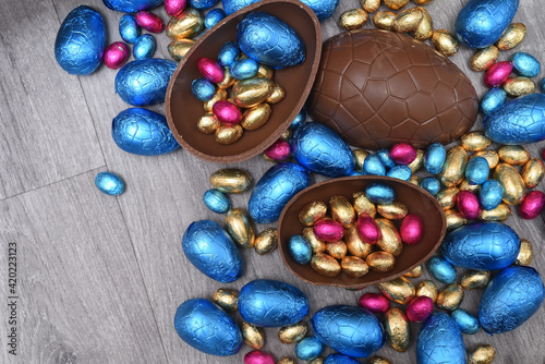Group of different sizes of colourful foil wrapped chocolate easter eggs in pink, blue, and gold. Large halves of a brown milk chocolate egg have mini eggs inside, on a grey wooden background.