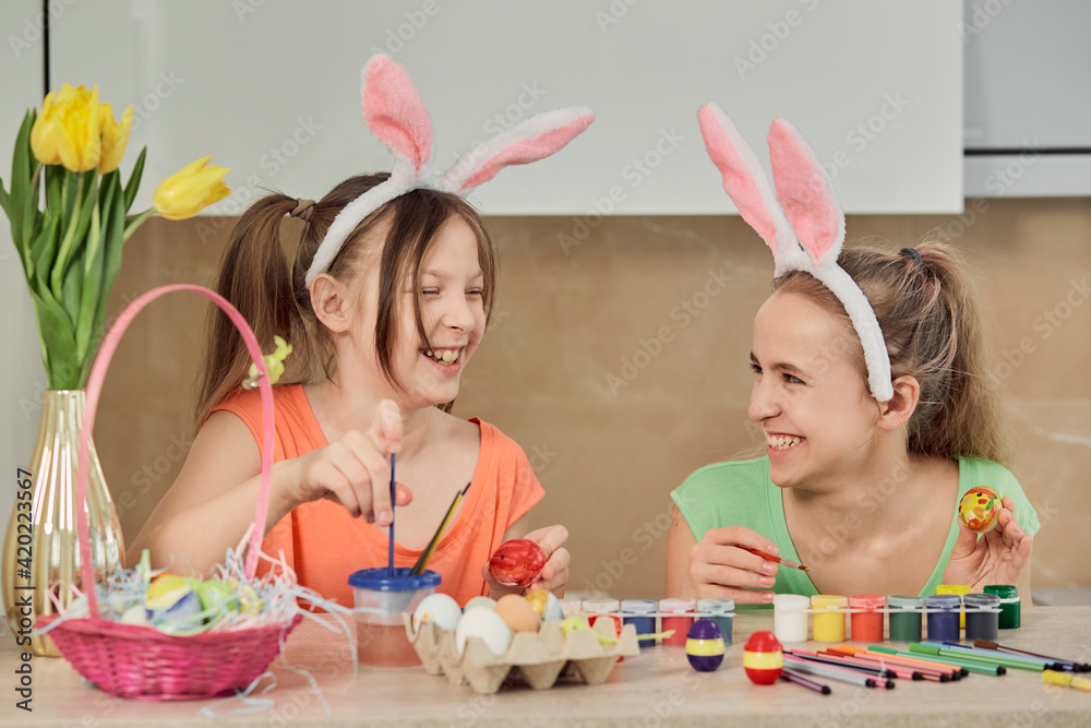 Two girls paint Easter eggs with colorful paint in the kitchen and laugh. Easter holiday. Two girls paint Easter eggs with colorful paint in the kitchen and laugh. Easter holiday.