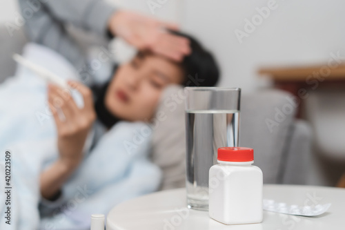 Bottle of medicine on the table and woman measuring temperature her body on the sofa.