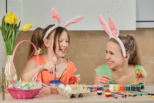 Two girls paint Easter eggs with colorful paint in the kitchen and laugh. Easter holiday.