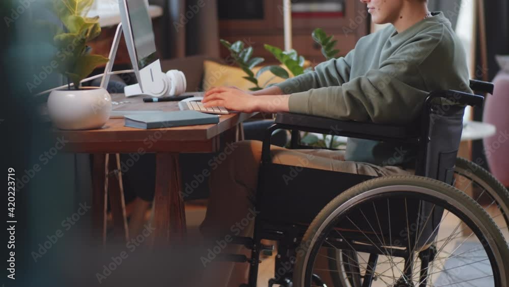 Midsection shot of young disabled woman on wheelchair typing on wireless computer keyboard at desk in living room at home