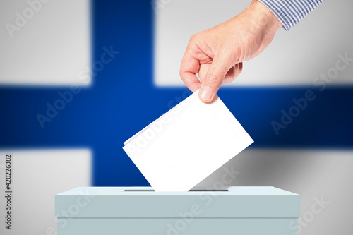 The hand of man putting his vote in the ballot box with Finland flag on background. Voting in election in Finland.
