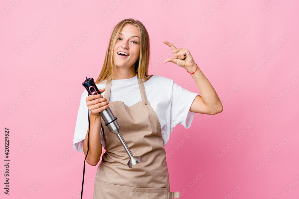 Young russian cook woman holding an electric mixer isolated holding something little with forefingers, smiling and confident. Young russian cook woman holding an electric mixer isolated holding something little with forefingers, smiling and confident.