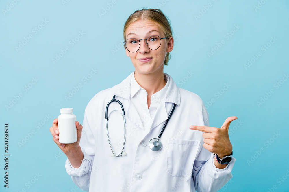 Young russian doctor woman holding pills bottle on blue person pointing by hand to a shirt copy space, proud and confident Young russian doctor woman holding pills bottle on blue person pointing by hand to a shirt copy space, proud and confident