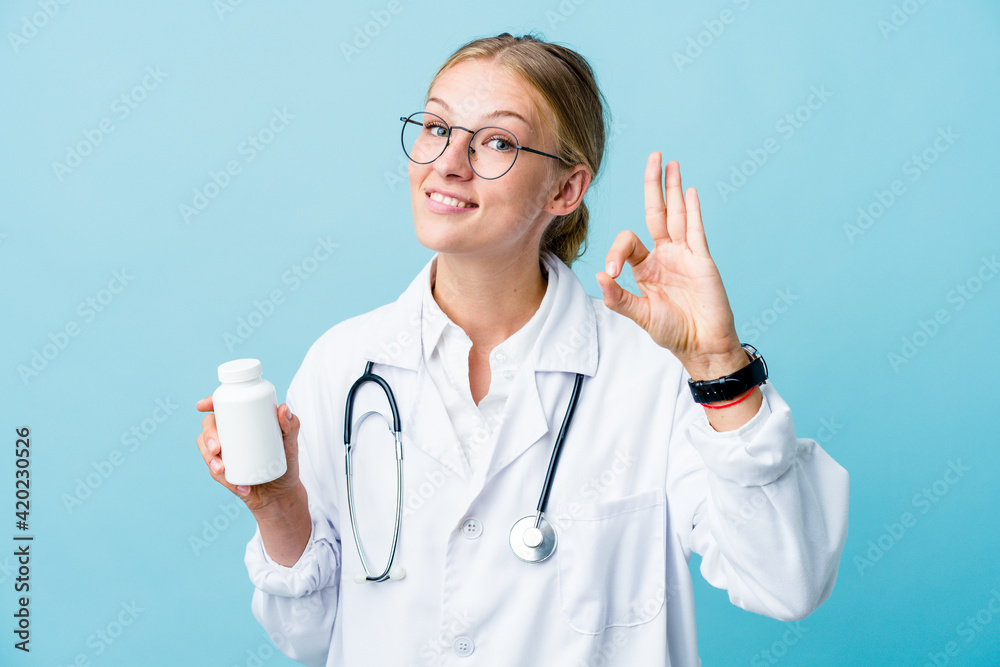 Young russian doctor woman holding pills bottle on blue cheerful and confident showing ok gesture. Young russian doctor woman holding pills bottle on blue cheerful and confident showing ok gesture.