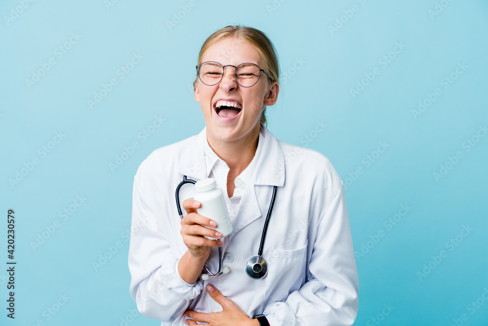 Young russian doctor woman holding pills bottle on blue laughs happily and has fun keeping hands on stomach. Young russian doctor woman holding pills bottle on blue laughs happily and has fun keeping hands on stomach.