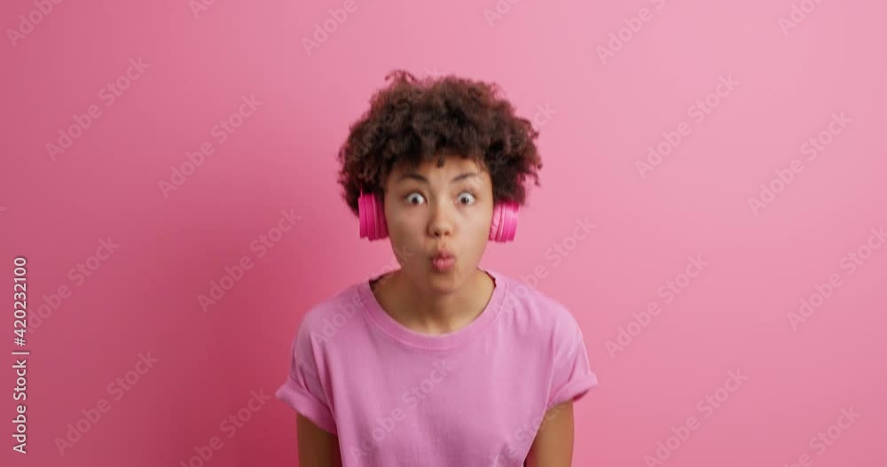 Positive carefree young African American teenage girl makes disco sign moves actively dances and shakes hands enjoys music in headphones dressed in casual t shirt isolated over pink background Positive carefree young African American teenage girl makes disco sign moves actively dances and shakes hands enjoys music in headphones dressed in casual t shirt isolated over pink background