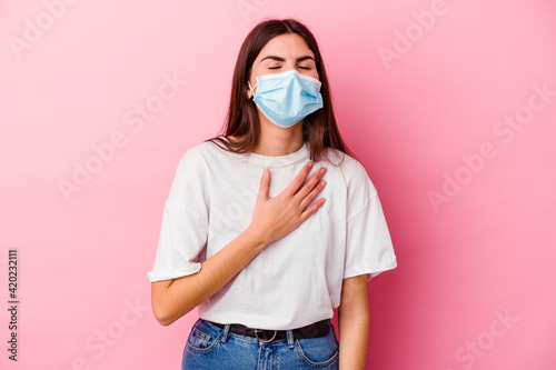 Young caucasian woman wearing a mask for virus isolated on pink background laughs out loudly keeping hand on chest.