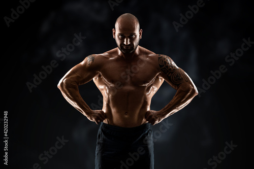 Male bodybuilder with light stubble and bare torso shows muscularity against a dark background. The concept of a fitness club, doing sports, weightlifting. Copy space.