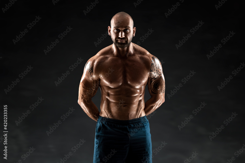 Fototapeta premium Male bodybuilder with light stubble and bare torso shows muscularity against a dark background. The concept of a fitness club, doing sports, weightlifting. Copy space.