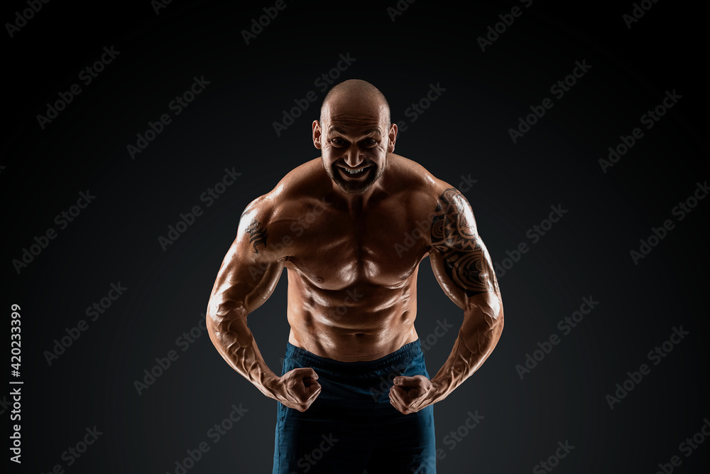 Fototapeta premium Male bodybuilder with light stubble with a naked torso posing against a dark background. The concept of a fitness club, doing sports, weightlifting. Copy space.
