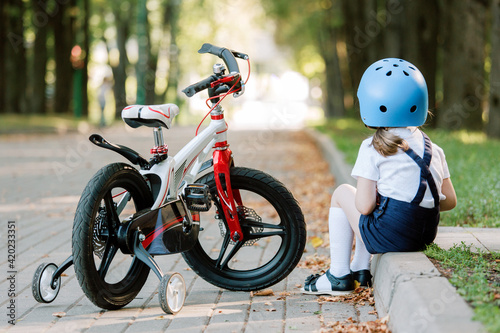 Backview of girl in helmet sitting near bicycle. Upset kid.