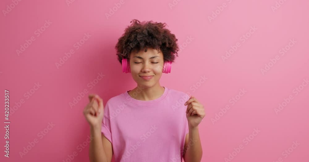 Sleepy tired African American woman yawns and moves slowly shakes hands dances carefree wears stereo headphones t shirt then smiles positively isolated over pink background. People lifestyle Sleepy tired African American woman yawns and moves slowly shakes hands dances carefree wears stereo headphones t shirt then smiles positively isolated over pink background. People lifestyle