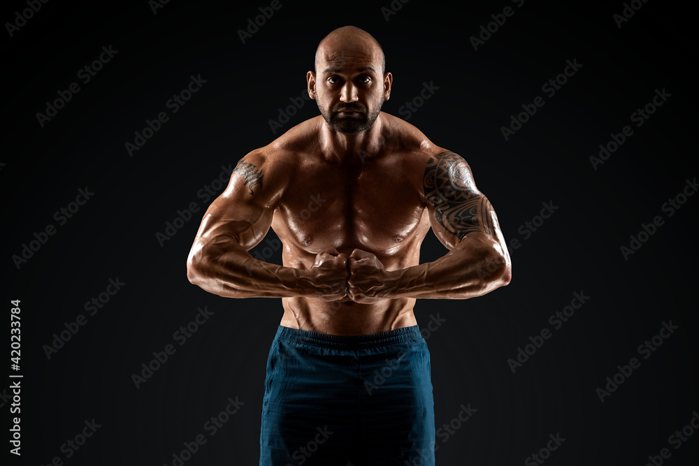 Fototapeta premium Male bodybuilder with light stubble with a naked torso posing against a dark background. The concept of a fitness club, doing sports, weightlifting. Copy space.