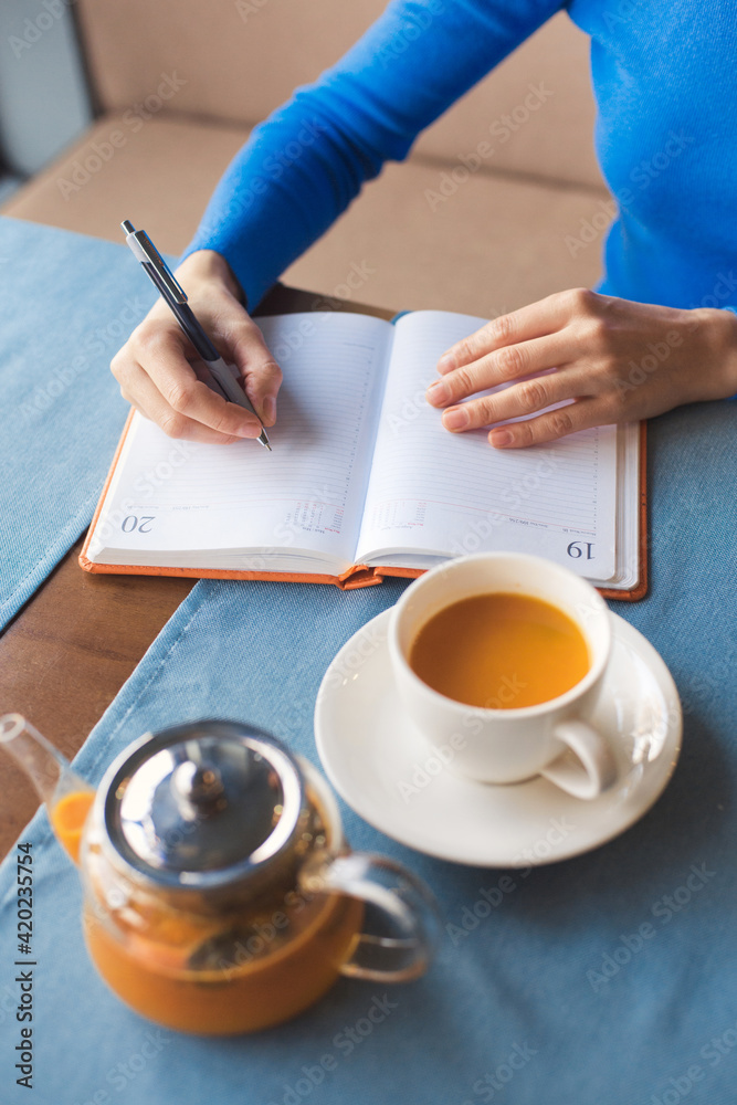 Woman have a teabreak during working hours and planning next work day. Girl writes her plans in a notebook and drinks vitaminic herbal healthy tea from orange and berry sea buckthorn, sitting in cafe Woman have a teabreak during working hours and planning next work day. Girl writes her plans in a notebook and drinks vitaminic herbal healthy tea from orange and berry sea buckthorn, sitting in cafe
