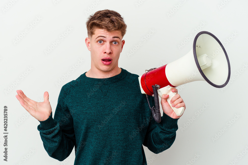 Young caucasian man holding a megaphone isolated on white background surprised and shocked. Young caucasian man holding a megaphone isolated on white background surprised and shocked.