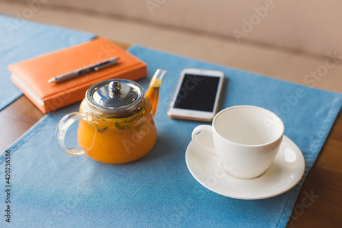 Someone have a teabreak during working hours and planning next work day. A cup of vitaminic herbal healthy tea from orange and berry sea buckthorn in glass teapot, notebook and phone. photo