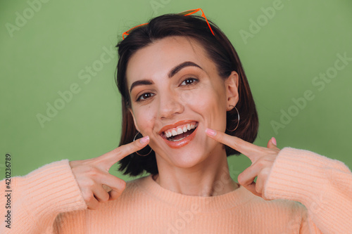 Young stylish woman in casual peach sweater and orange glasses isolated on green olive background positive smiling pointing on white teeth with index fingers copy space