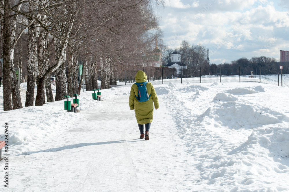 A woman in a yellow long coat with a backpack walks along the park alley on a winter day.