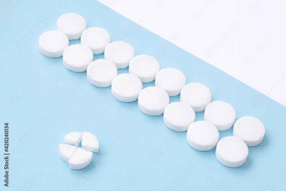 Round white medical pills arranged and one of them is crushed into four parts on blue background. Pill splitting.