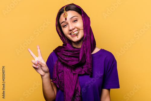 Young Indian woman wearing a traditional sari clothes isolated on yellow background joyful and carefree showing a peace symbol with fingers.