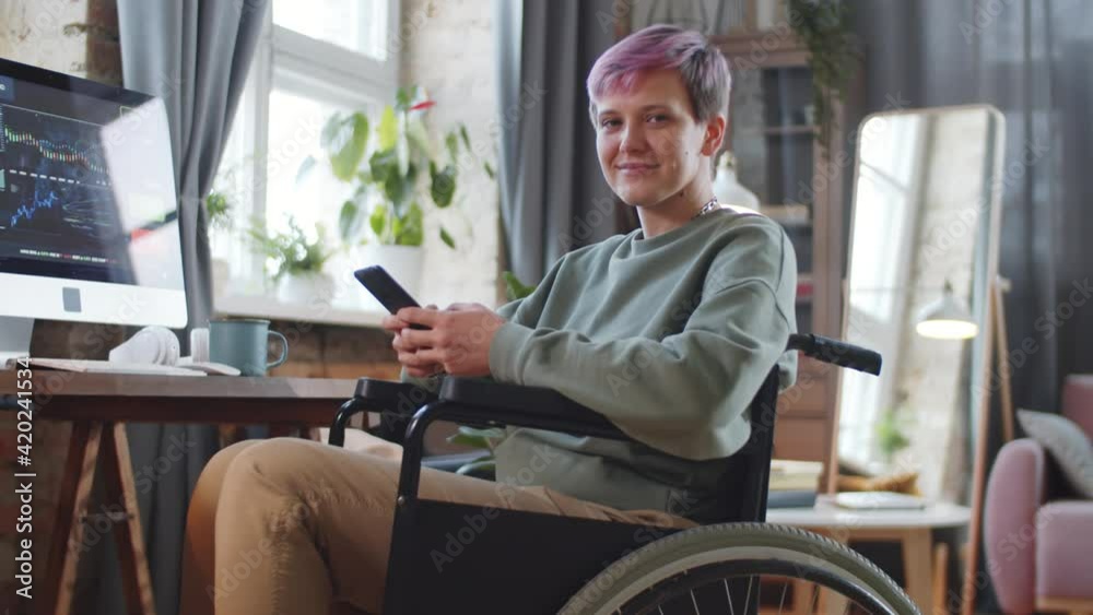 Young cheerful disabled woman on wheelchair holding smartphone, looking at camera and smiling while working remotely from home