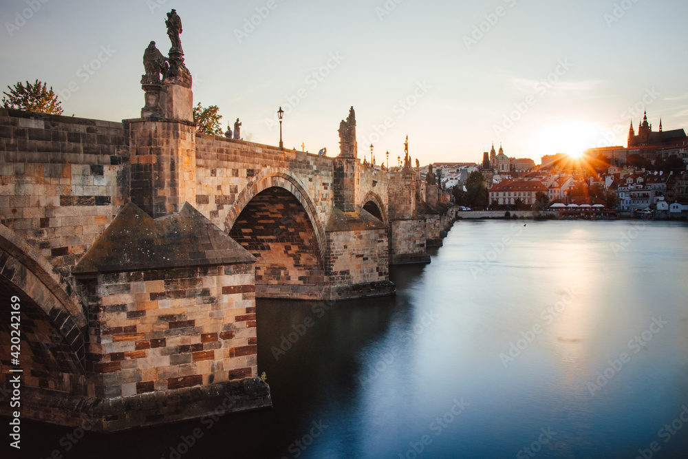 Obraz premium famous stone bridge in Prague in the Czech Republic known as Charles Bridge connects parts of the city across the Vltava River at sunset. Long exposure of river, sun and Charles Bridge