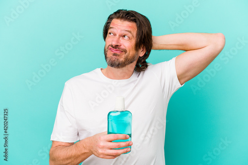 Middle age dutch man sitting holding a mouthwash isolated on blue background touching back of head, thinking and making a choice. © Asier