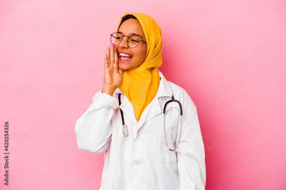Young doctor muslim woman isolated on pink background shouting and holding palm near opened mouth. Young doctor muslim woman isolated on pink background shouting and holding palm near opened mouth.