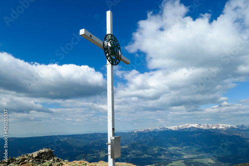 A metal cross on the top of Ameringkogel in Austrian Alps. The cross is surrounded by piles of stones. There are other chains in the back. Blue sky with white clouds on it. Serenity and achievement.