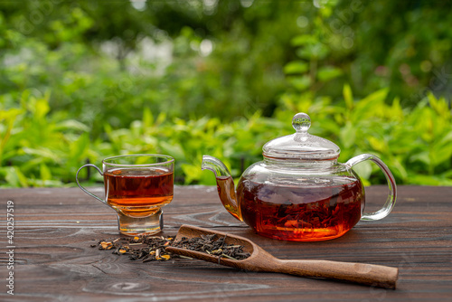 Steamed glass teapot, elegant cup. Bamboo spoon with tea, wooden table. Outdoor, picnic, brunch. Floral background in blur.