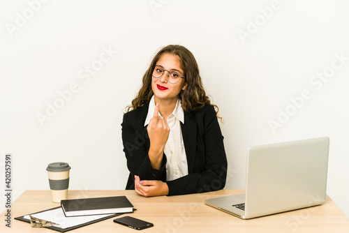 Young caucasian business woman working on her desktop isolated pointing with finger at you as if inviting come closer.