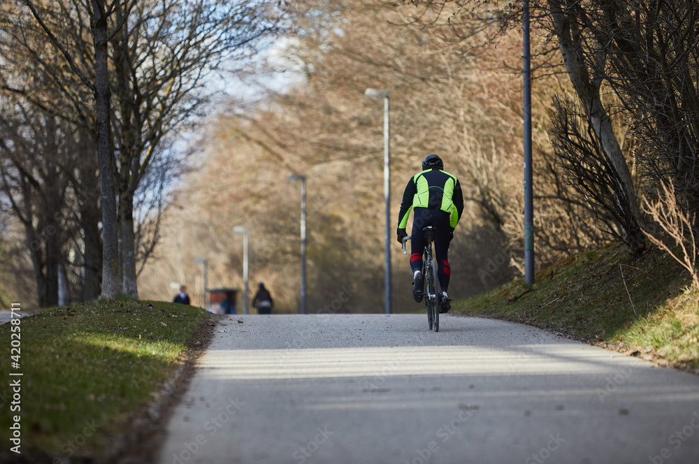 Man riding a road bike. You see him from behind as he pedals standing. The sun is shining and you see a bicycle road.