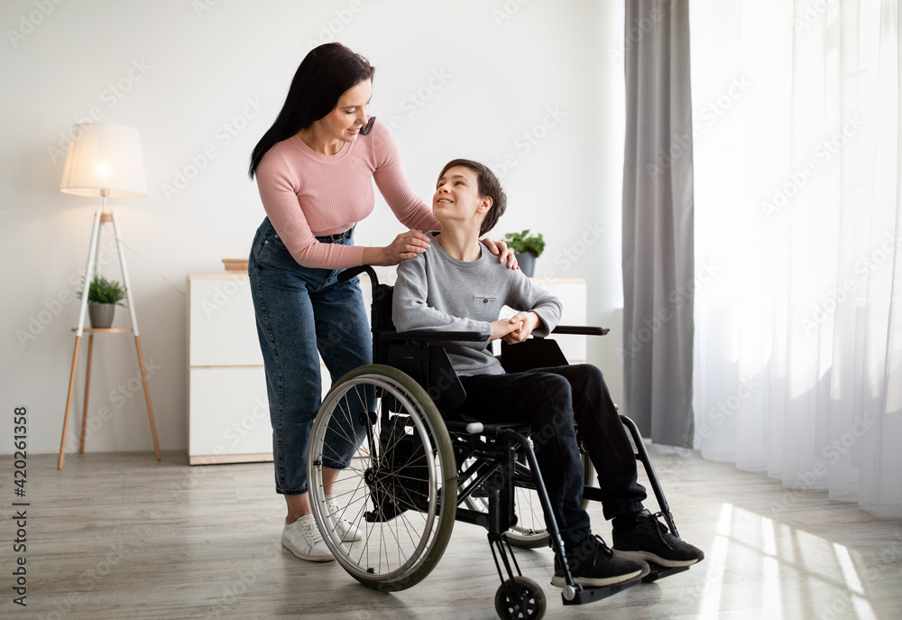 Full length portrait of mother and her disabled son in wheelchair looking at each other and smiling at home Full length portrait of mother and her disabled son in wheelchair looking at each other and smiling at home